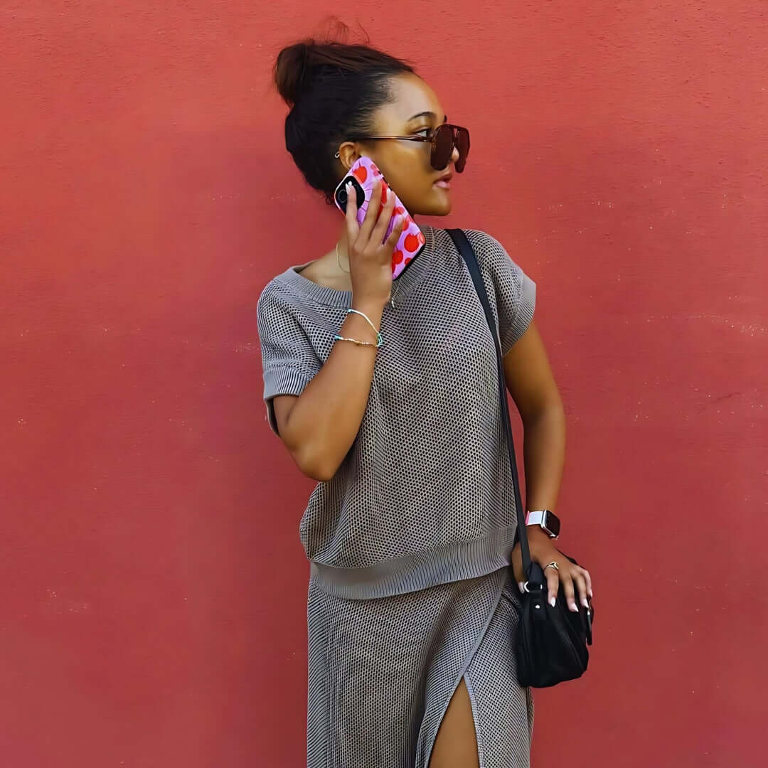 Woman holding iPhone 14 with cute phone case, dressed in grey outfit, standing against red wall.